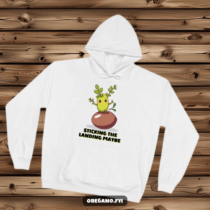 Funny hoodie showing an oregano leaf character attempting a shaky balance on a wobbly olive pit, offering cozy amusement.